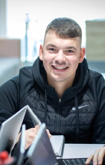 Smiling male student in a black hoodie working with classmates on laptops at a study area in Glasgow Kelvin College’s Springburn Campus. Smiling male student in a black hoodie working with classmates on laptops at a study area in Glasgow Kelvin College’s Springburn Campus.